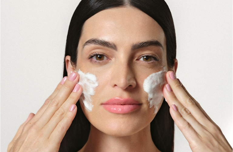 Foto de una mujer, con fondo claro, aplicándose crema en el rostro con ambas manos, con espuma del producto visible en las mejillas.
